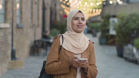Muslim-Woman-Wearing-Hijab-Walking-Along-City-Street-Sending-Text-Message-To-Date-On-Mobile-Phone